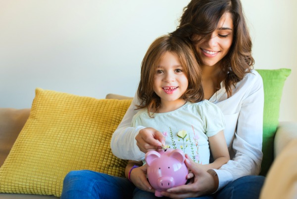 brunette mother sitting on a couch with a young girl on her lap putting money into a piggy bank