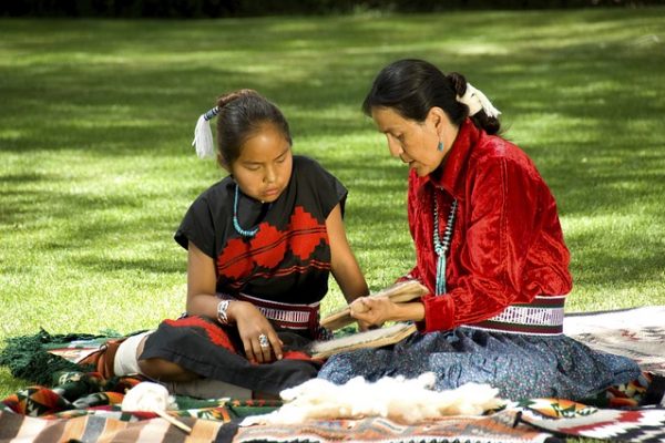 Native American Heritage Month Lessons + Make a Totem Pole Craft! an indigenous grandmother and granddaughter sitting on the ground working on a craft