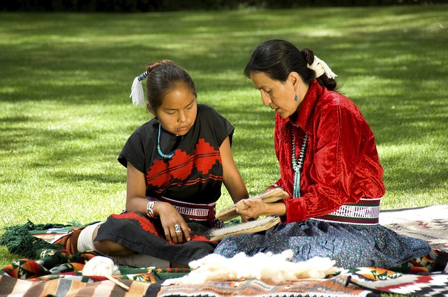 Native American Heritage Month Lessons + Make a Totem Pole Craft! an indigenous grandmother and granddaughter sitting on the ground working on a craft