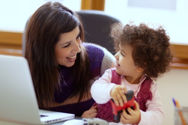 young mom with a laptop on a table and smiling at a toddler sitting next to her