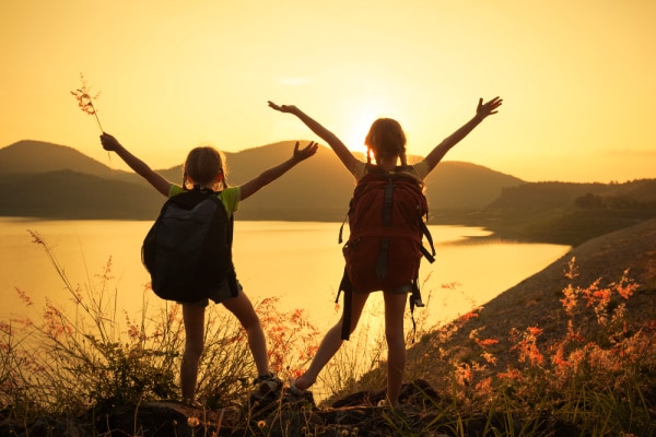 How To Hike With Kids two tween girls with hiking backpacks looking at water with arms up