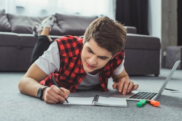 Remote Learning Tips for Parents and students caucasian teen male lying on stomach on floor working on laptop and writing in notebook for a remote learning class